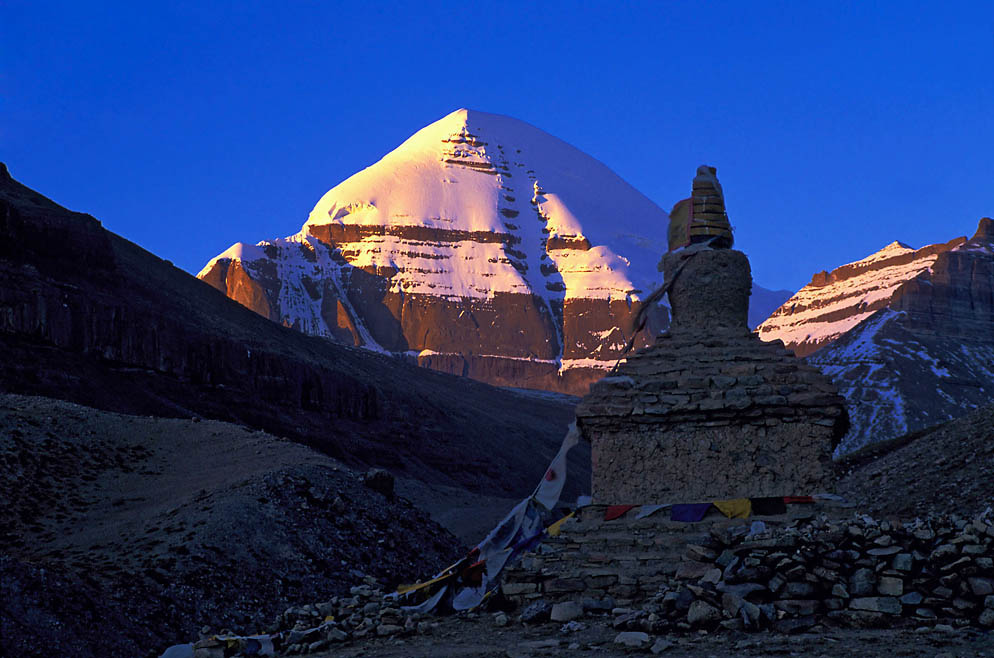 A serene scene of Mount Kailash at sunset, its snow-capped peak glowing softly under a deep blue sky. In the foreground, a white stupa stands quietly among rugged terrain, symbolizing peace and spiritual reverence in the vast, sacred landscape.